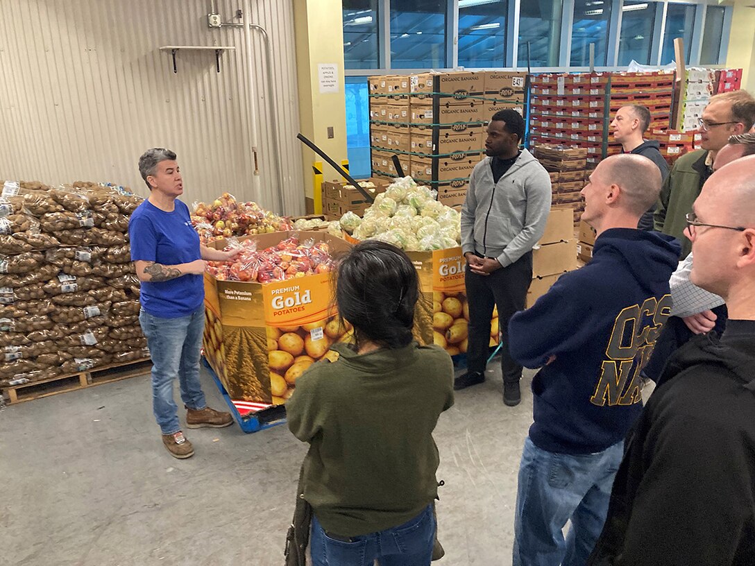Several people stand around in a warehouse full of food listening to a woman in a blue T-Shirt and jeans with graying hair speak.