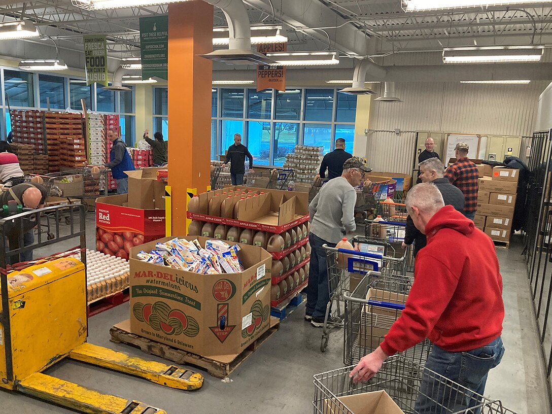 Several people wearing casual clothing load shopping carts with food in a warehouse.