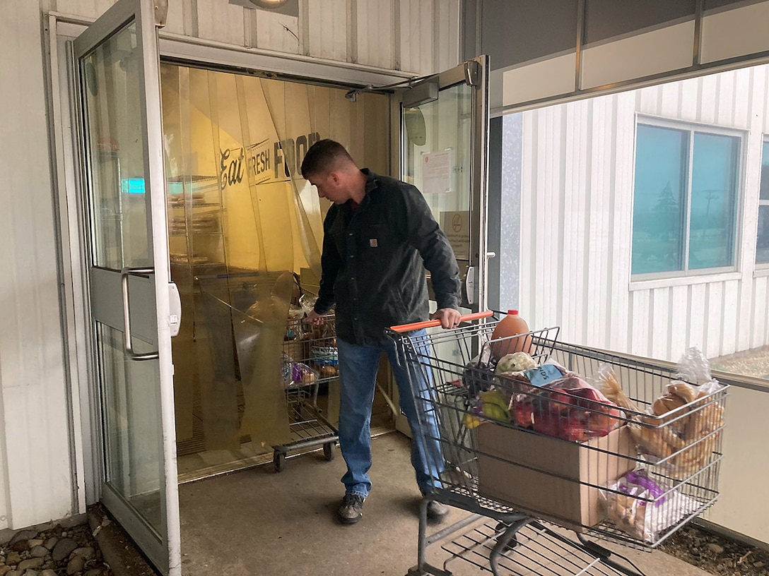 A man in a a dark shirt and jeans wearing pushes a shopping cart full of food.