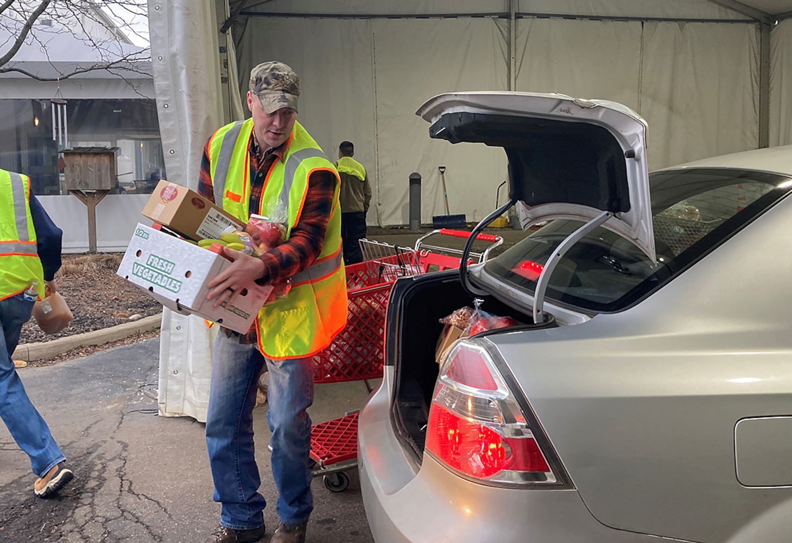 A man in a a yellow reflective vest and jeans wearing a ballcap loads a car trunk with food.