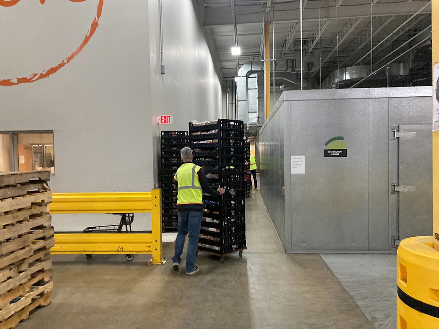 A man in a yellow reflective vest and jeans with graying hair moves a tower of pallets in a warehouse.