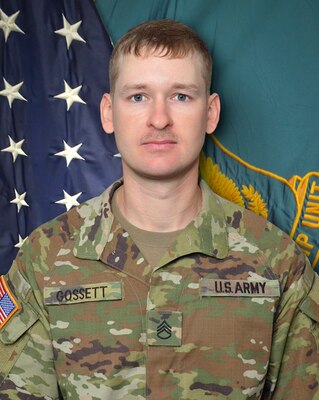 Man in U.S. Army uniform standing in front of two flags.