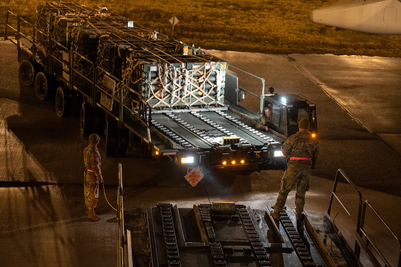 Pallets of equipment are loaded onto large vehicles on a flight line.