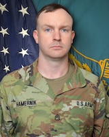 Man in U.S. Army uniform standing in front of two flags.