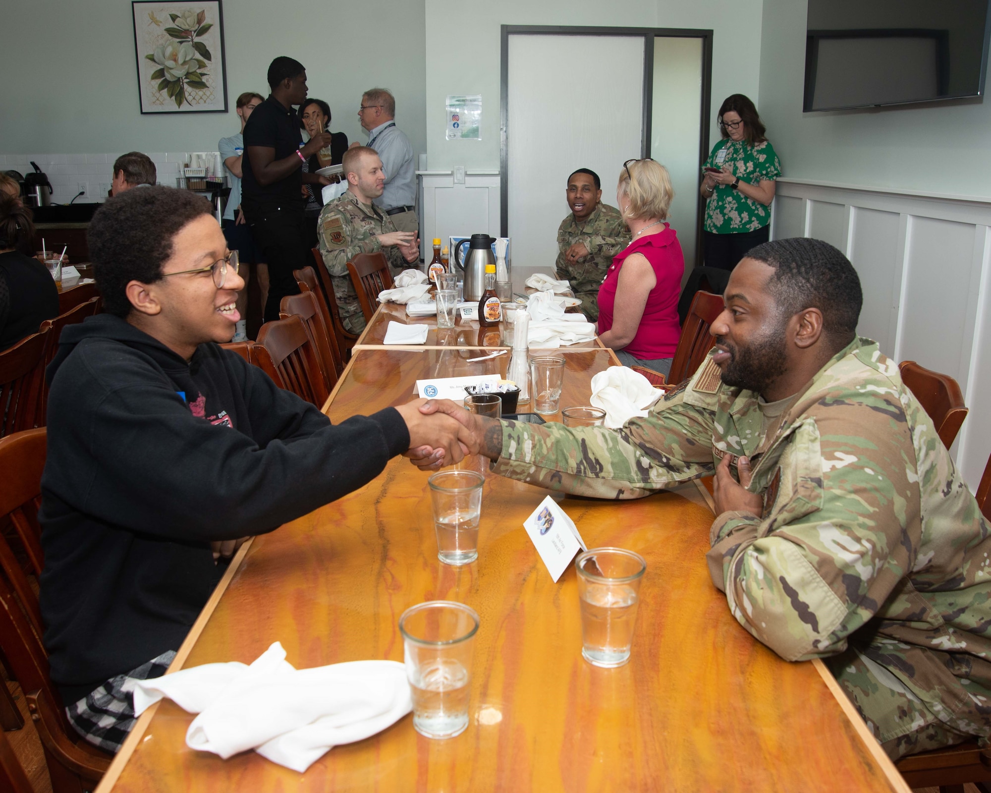 A person in a camouflage uniform shakes hands with a student while sitting at a table.