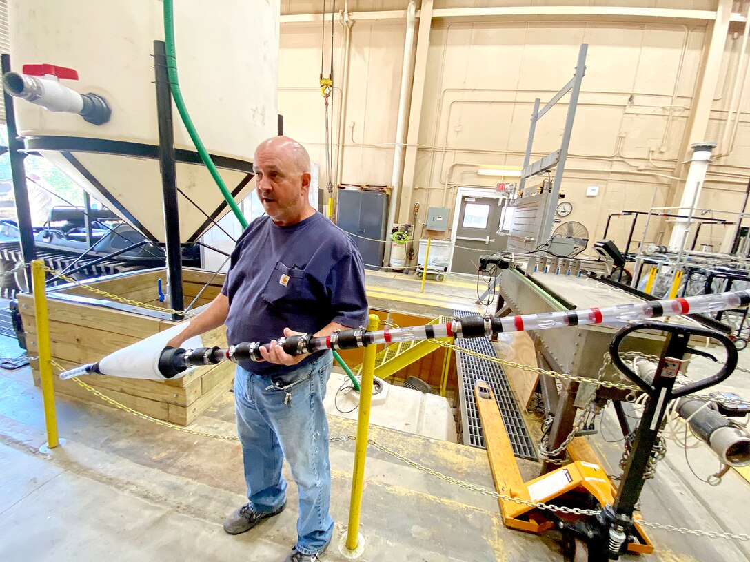 A man wearing a blue shirt and jeans stands in warehouse holding a piece of equipment.