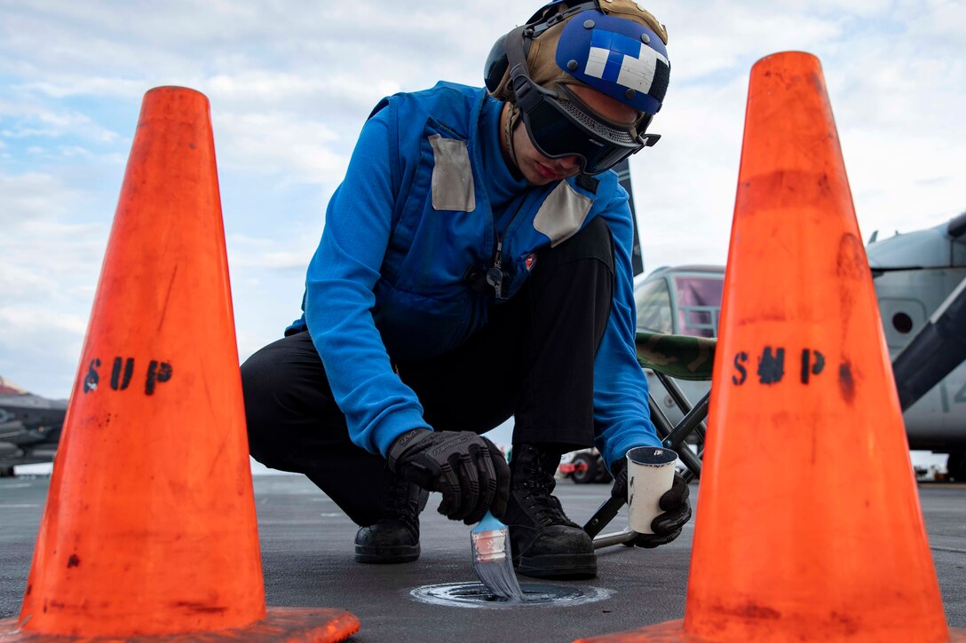 A sailor in blue wearing helmet and goggles uses a brush to paint a padeye between two large orange cones on the deck of a ship.