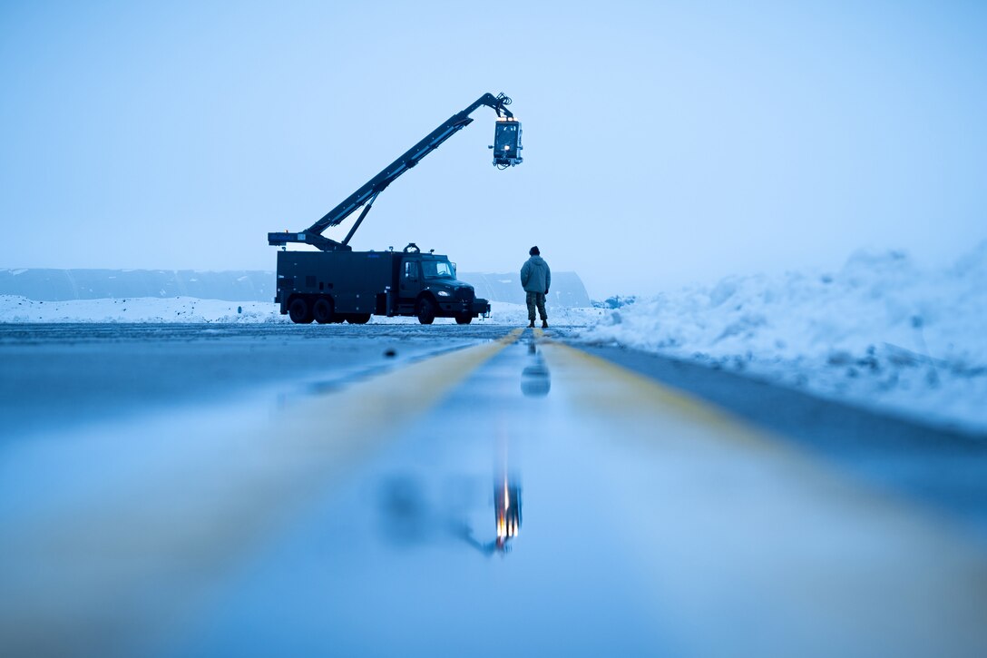 An airman watches as a large truck with a crane de-ices a runway.