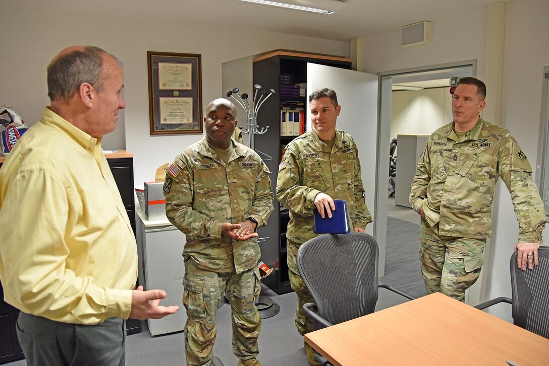 Four men stand near a desk talking.