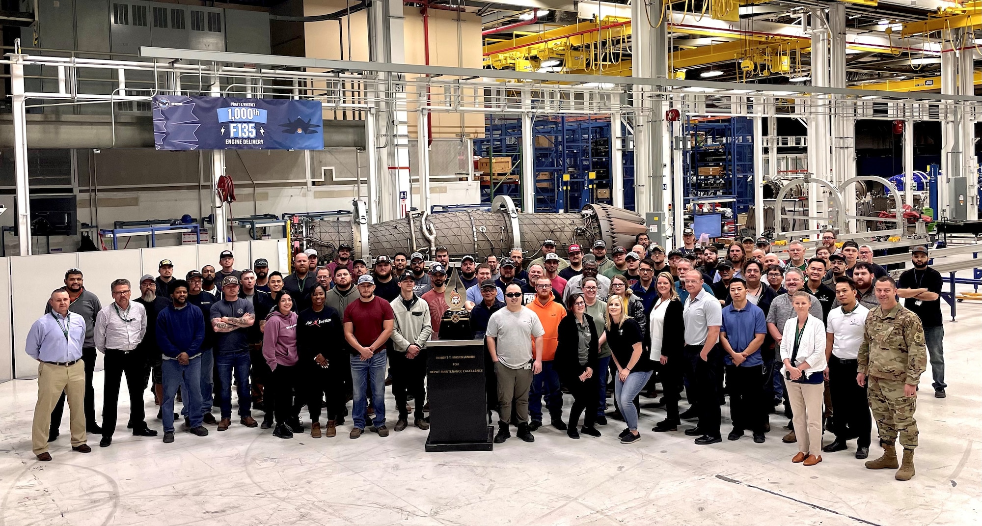 People posing for group photo with award in foreground and jet engine in background.