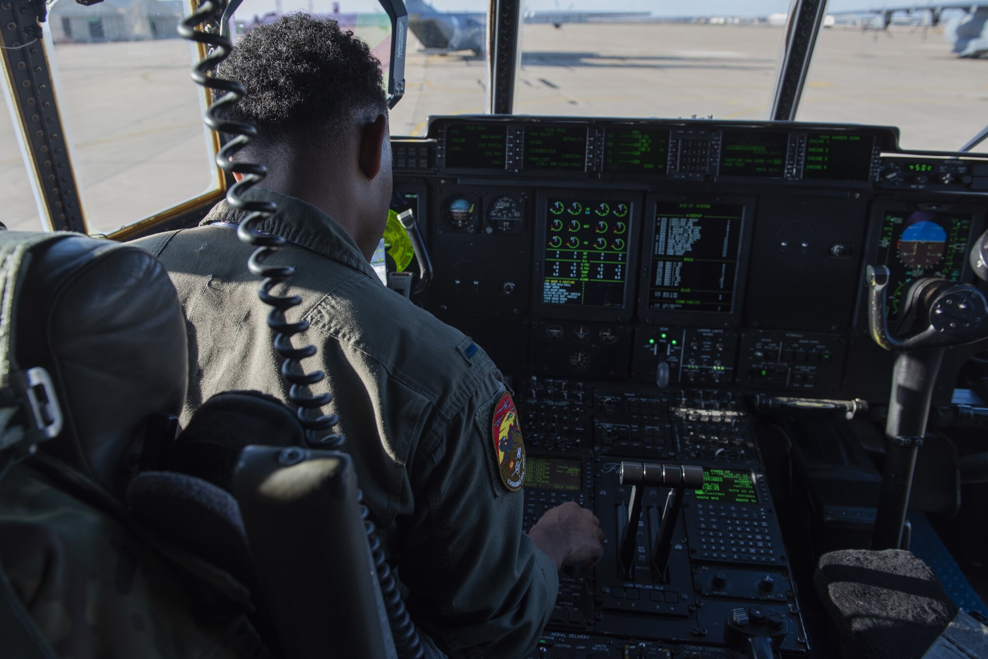 A pilot in a green suit sits in a seat in the cockpit of a plane and presses a button