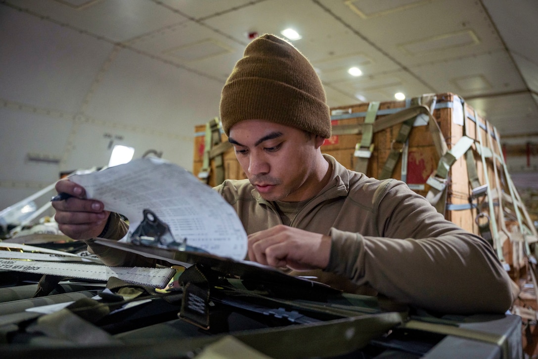 An airman checks cargo logs attached to a clipboard with cargo in the background.