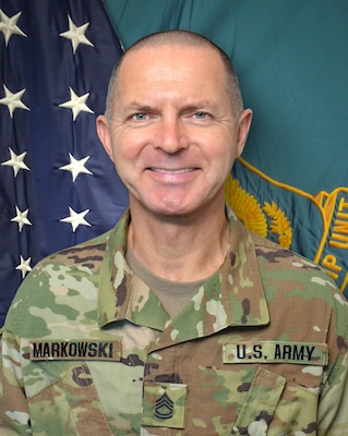 Man in U.S. Army uniform standing in front of two flags.