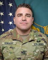 Man in U.S. Army uniform standing in front of two flags.