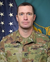 Man in U.S. Army uniform standing in front of two flags.