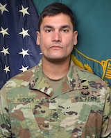 Man in U.S. Army uniform standing in front of two flags.