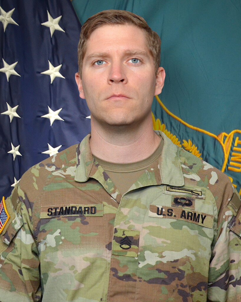Man in U.S. Army uniform standing in front of two flags.