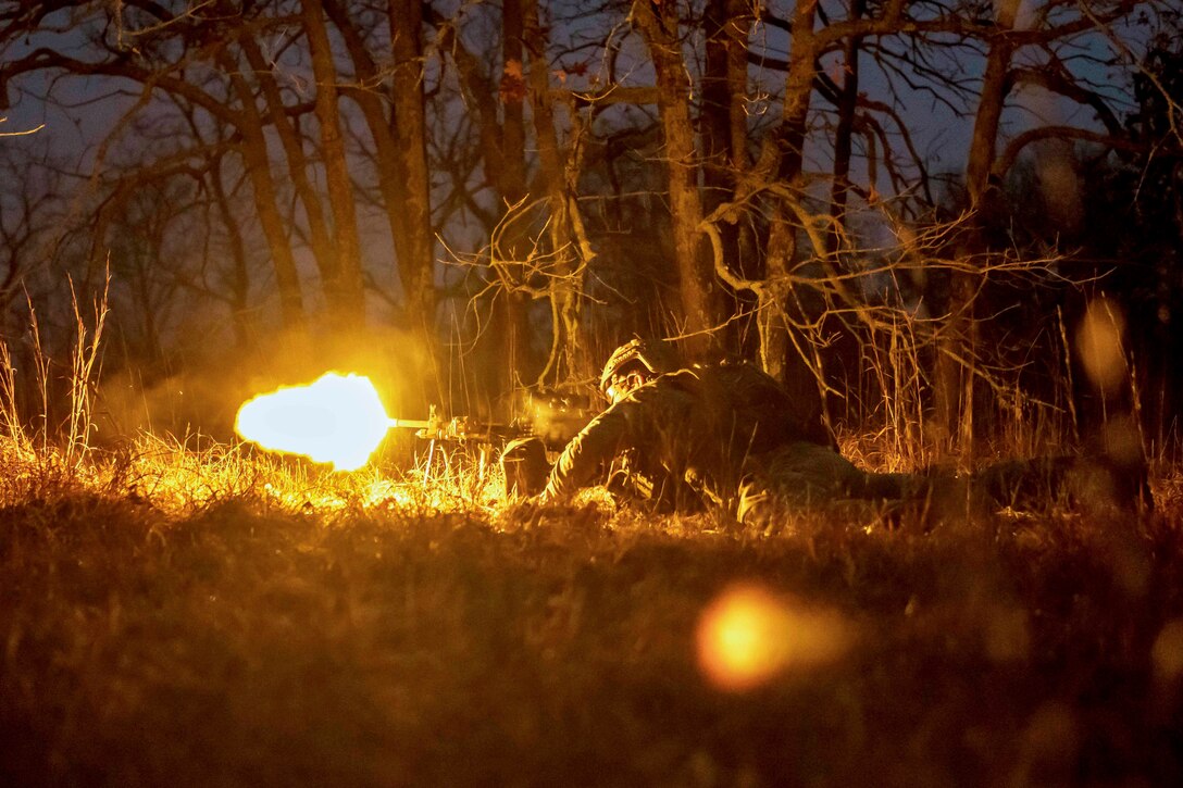 A sailor laying on the ground in the woods fires a weapon in the dark illuminated by gun fire.