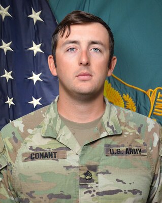 Man in U.S. Army uniform standing in front of two flags.