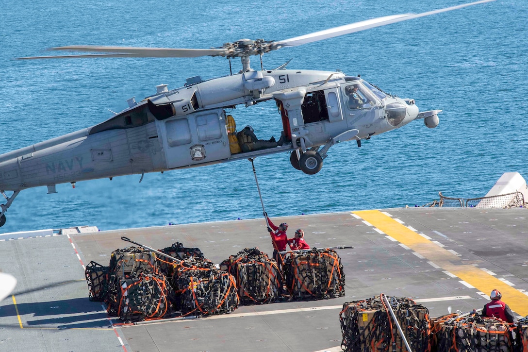 Two sailors standing amid pallets on a ship's flight deck attach a hook to a hovering helicopter.