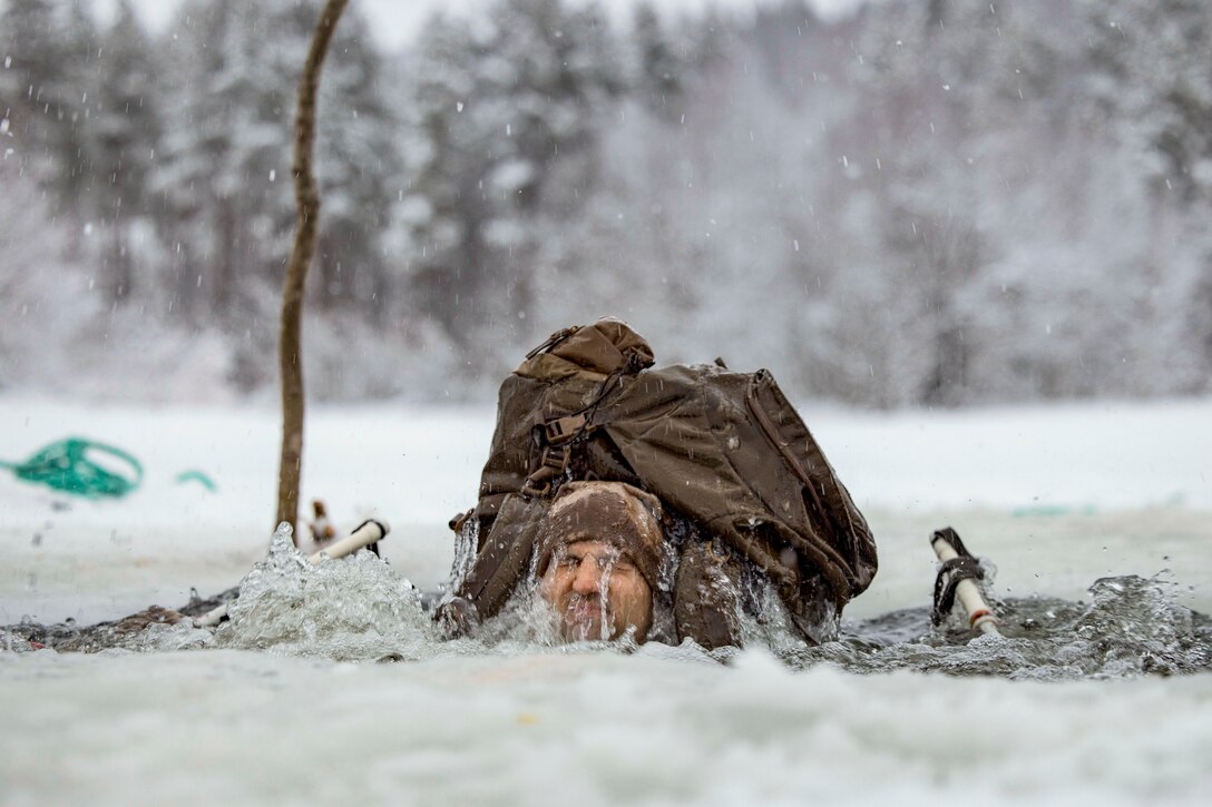 A Marine wearing a backpack grimaces while submerged in water surrounded by snowy terrain.