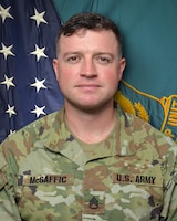 Man in U.S. Army uniform standing in front of two flags.