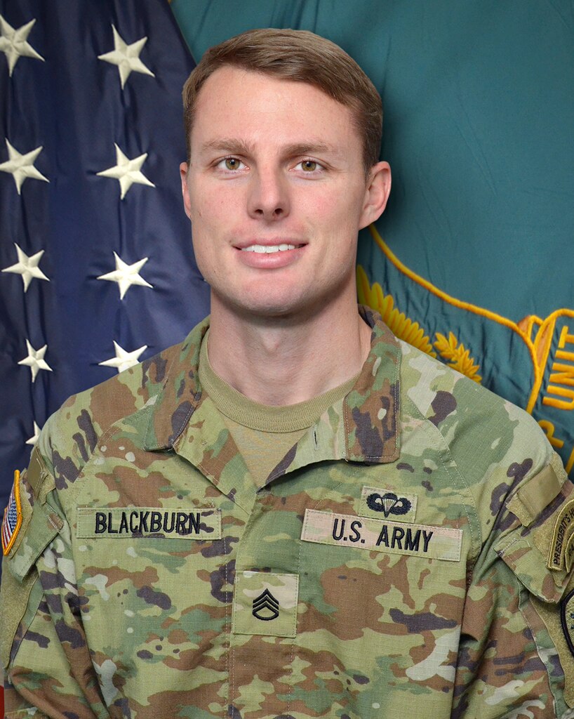 Man in U.S. Army uniform standing in front of two flags.