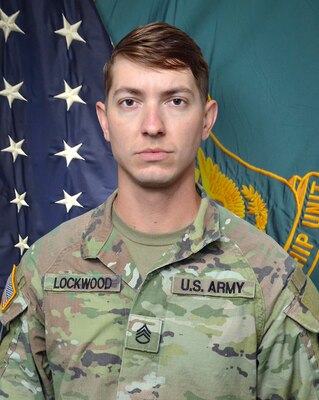 Man in U.S. Army uniform standing in front of two flags.