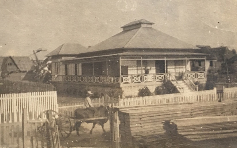 A pile of lumber sits by a dirt road in front of a large house.