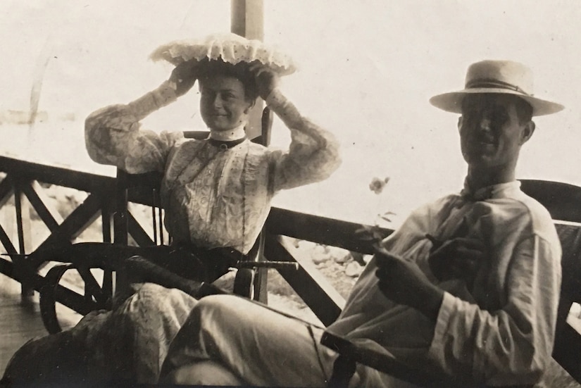 A man and woman smile while sitting on chairs on a porch. The man’s arm is in a sling.