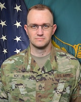 Man in U.S. Army uniform standing in front of two flags.