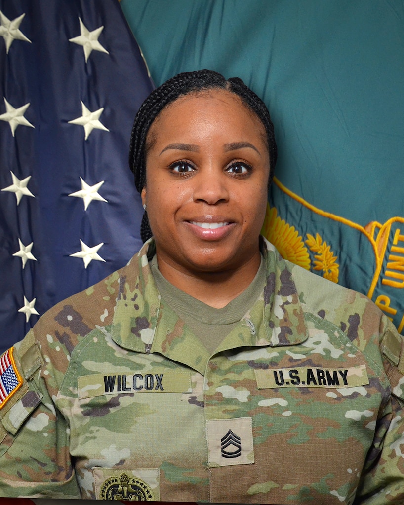 Woman in U.S. Army uniform standing in front of two flags.