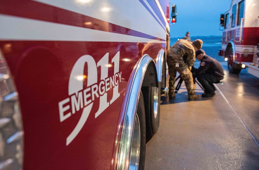 An airman and a civilian kneel to inspect a light next to a fire engine.