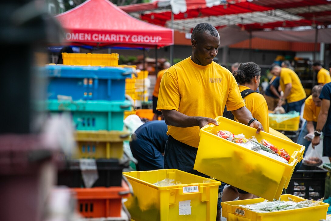 A sailor sorts through bins of food  in a crowded area.