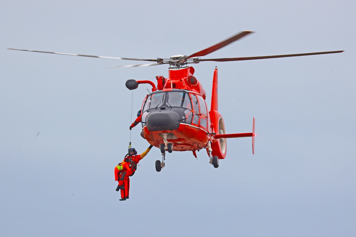 Orange helicopter hovering mid-air while two uniformed coast guardsmen hang from hoist attached to helicopter.
