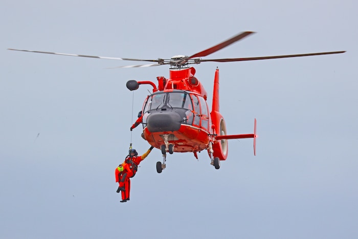 Orange helicopter hovering mid-air while two uniformed coast guardsmen hang from hoist attached to helicopter.