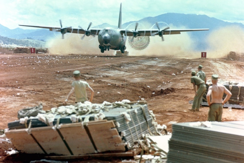 Service members stand in a dusty field as an airplane taxis