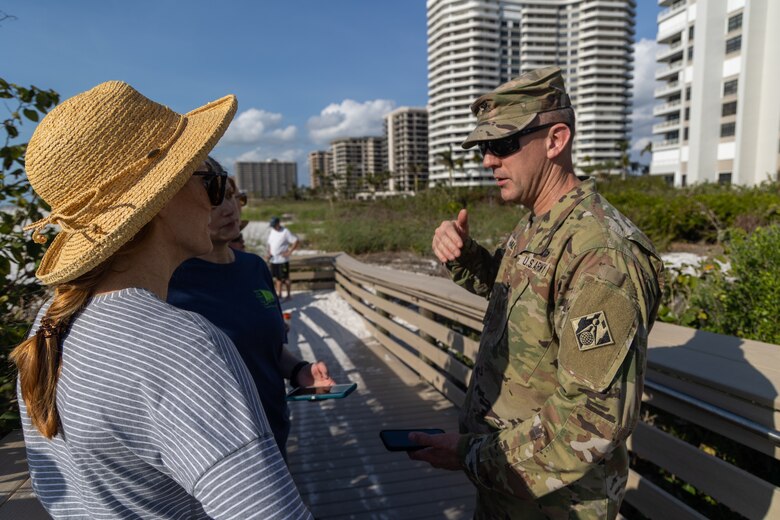 Col. Brian Hallberg stands on a wood ramp in uniform on the right hand side of the image, He is talking a woman on in a stripped long sleeve shirt wearing a sun hat, and another woman in a dark blue t-shirt. Condos and tropical vegetation are in the background
