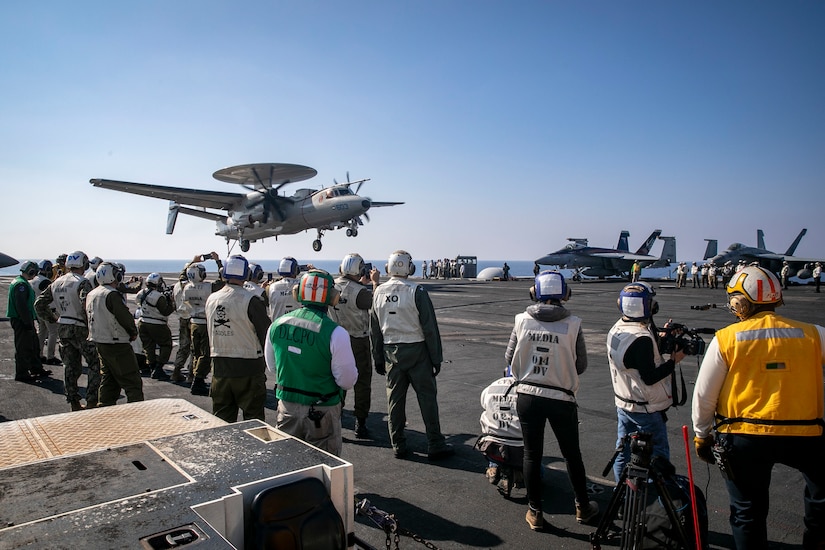 An aircraft lands on the deck of a large ship. Dozens of people stand nearby.