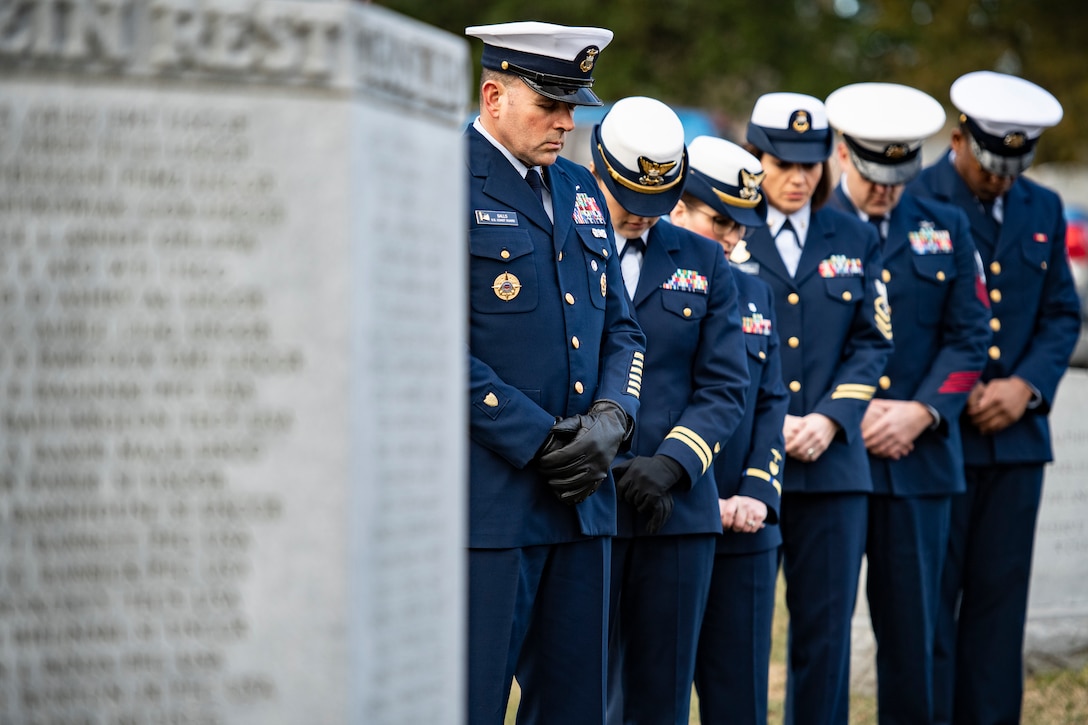 A group of Coast Guardsmen stand and  bow their heads next to a memorial.