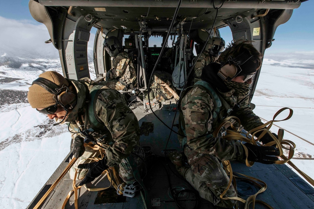 Two soldiers pull in lines from inside an airborne helicopter.