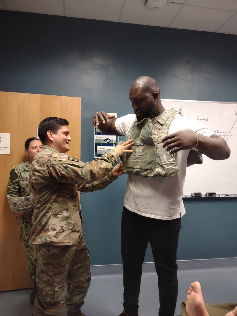 Staff Sgt. Jose Diaz, an instructor in the Medical Education and Training Campus Respiratory Therapist program, fits Omos, one of the WWE wrestling Superstars who visited Joint Base San Antonio-Fort Sam Houston on Jan. 26, with an improved outer tactical vest that is worn on deployment, even when conducting patient care.