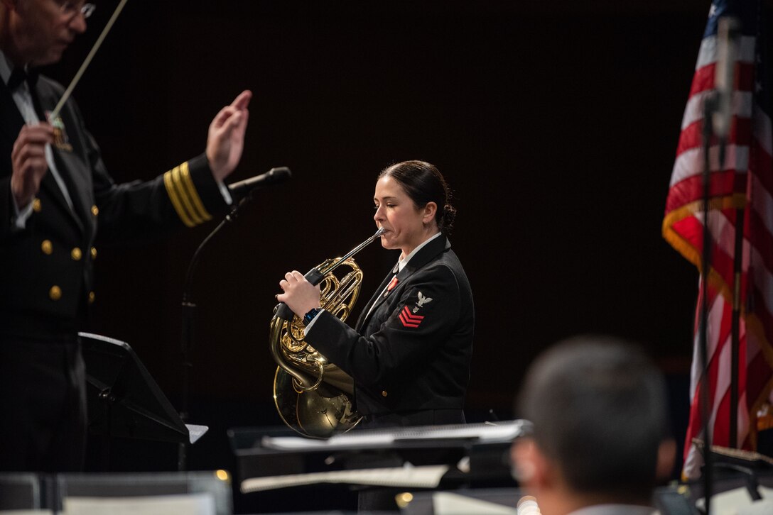 A sailor conducts while another plays the horn during a band performance with the American flag off to the side.