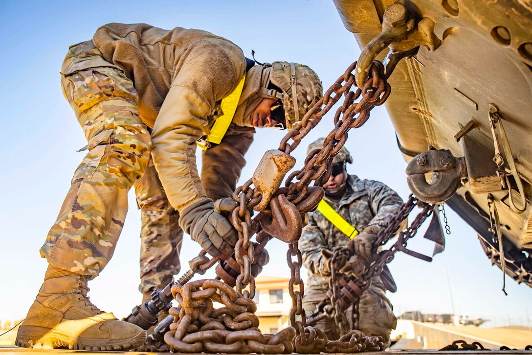 Two soldiers tighten chains on a military vehicle.