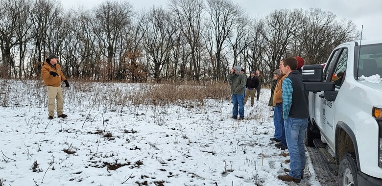 People stand outside in a snowy field with trees listening to a person discuss natural resources.