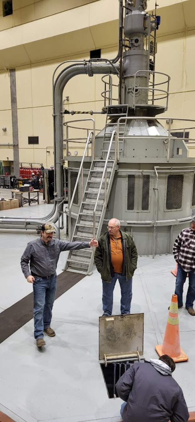 Men stand in a room with a big metal hydropower turbine behind them as they talk.