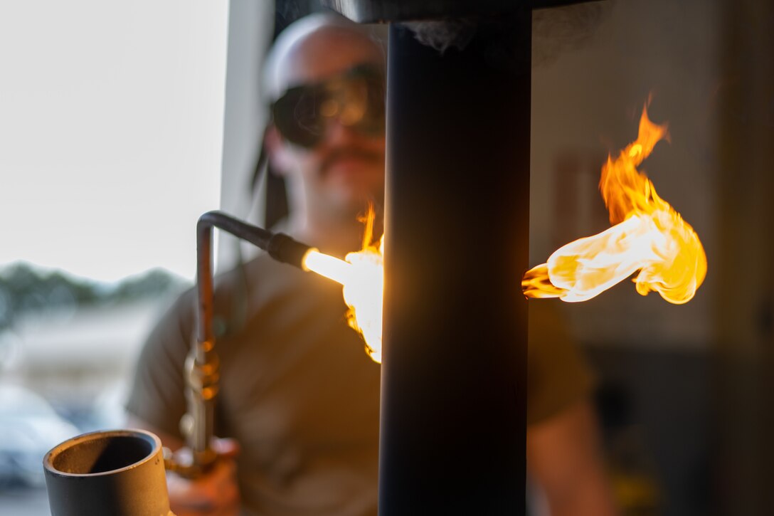 A man wearing goggles is shown close-up, operating a blow torch.