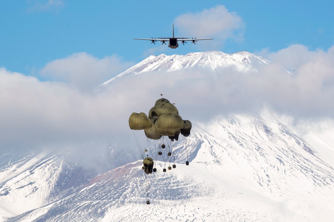 Containers free fall with parachutes over snowy mountains as an aircraft flies above.