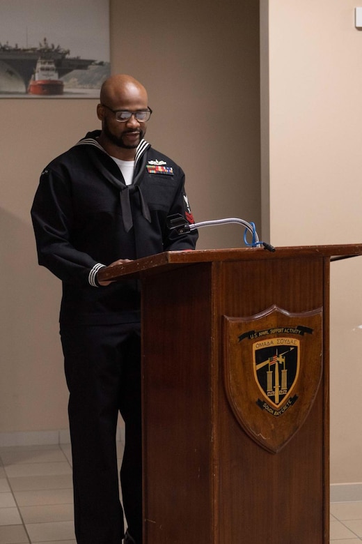 A man in a Navy dress uniform stands at a lectern.