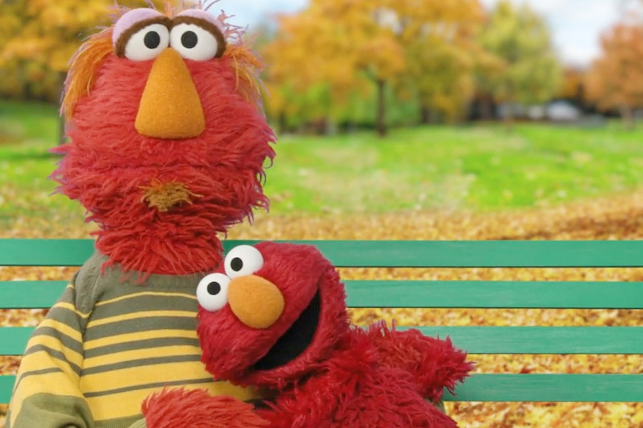 A little puppet rests its head on the shoulder of a large puppet as they sit on a park bench.